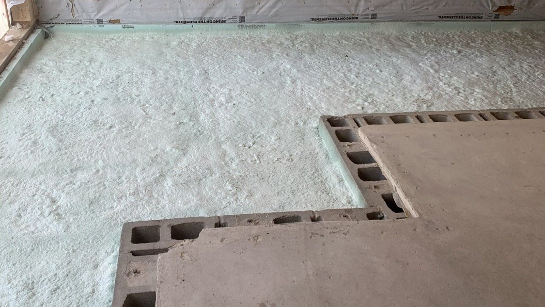 Below slab insulation on a basement floor: closed-cell spray foam applied over the base before the pour, with board along the wall and the old slab beside it