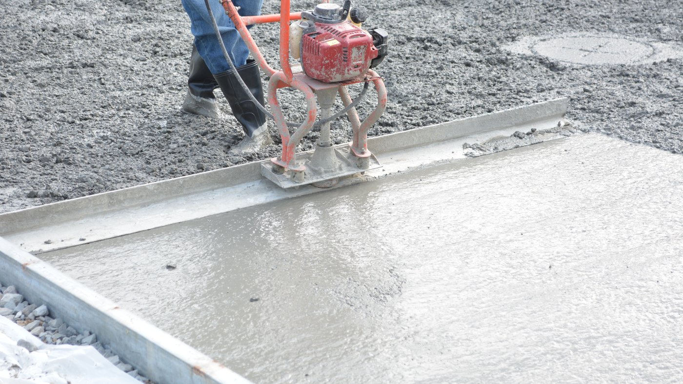 Fresh concrete being screeded level with a power screed, the gravel base and polyethylene sheet still showing at the edge of the pour
