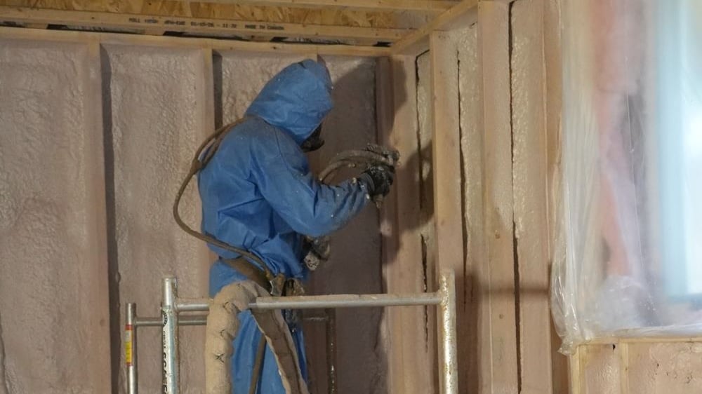 Applicator in a full protective suit spraying closed cell foam into the stud bays of a framed wall