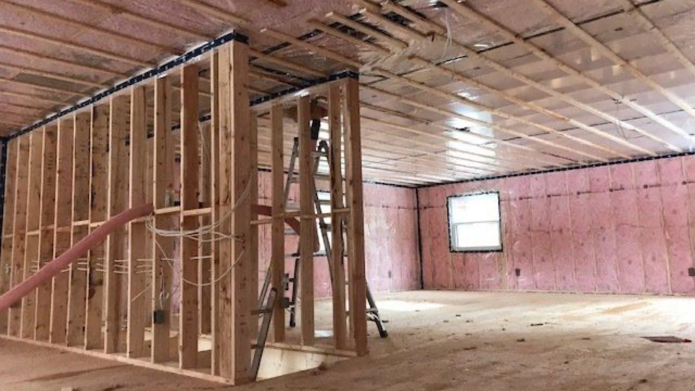 New-build interior before drywall: pink batts in the exterior walls behind clear poly taped at the top plates, poly on the ceiling, and a framed interior wall with wiring in the foreground
