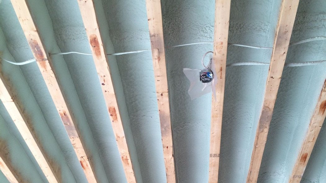 Garage ceiling joists with closed-cell spray foam tight to the sheathing above, the way floor insulation over a garage is sprayed, with wiring run across the bays