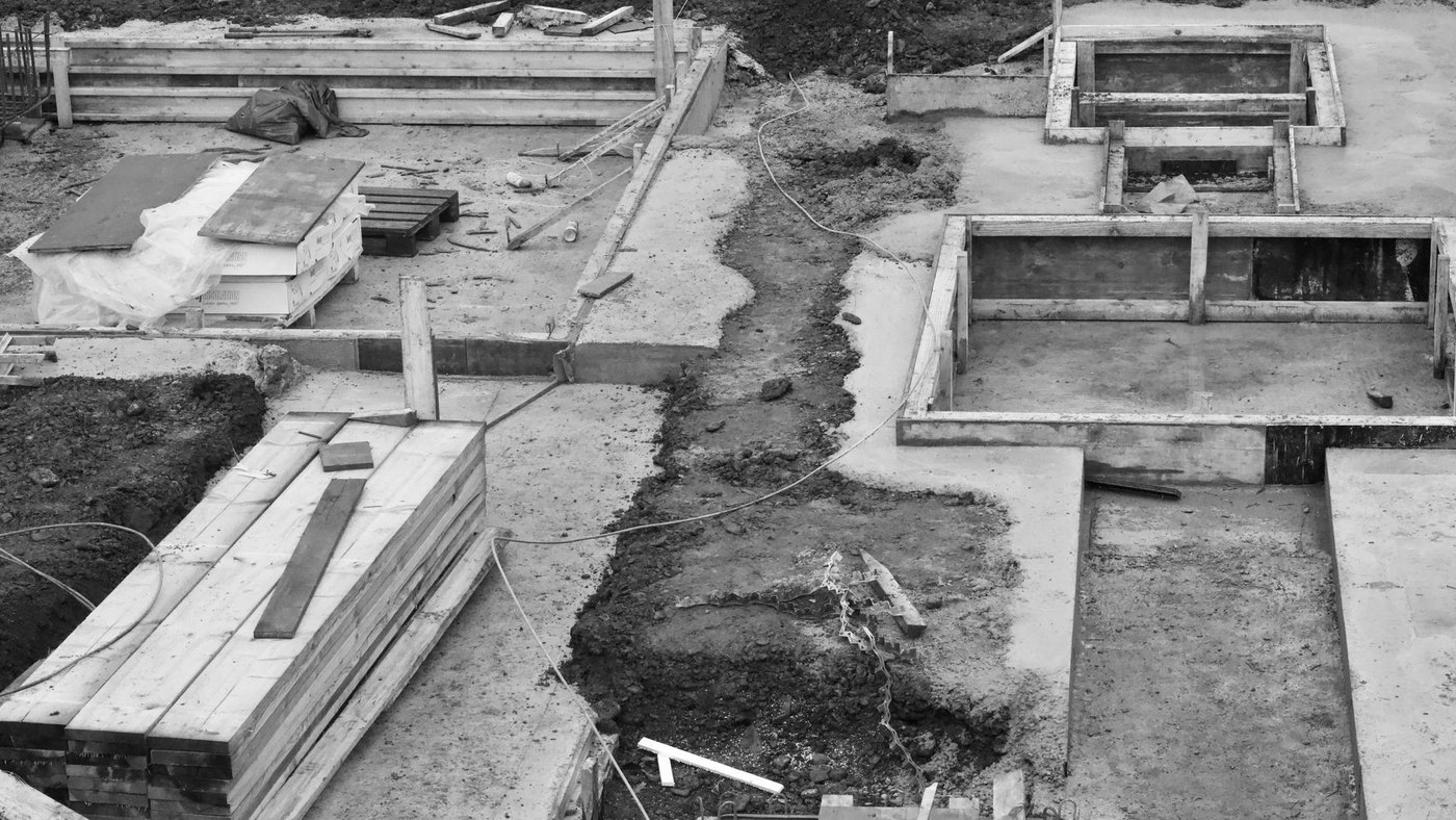 Frost wall footings and short foundation walls formed on an open site before backfill, with wrapped packs of board stock on a pallet beside them