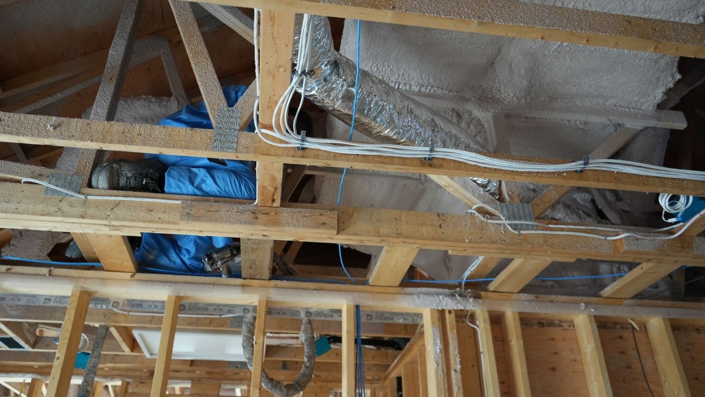 Applicator in a blue protective suit spraying closed-cell foam onto the roof deck above the ductwork and wiring of a framed house