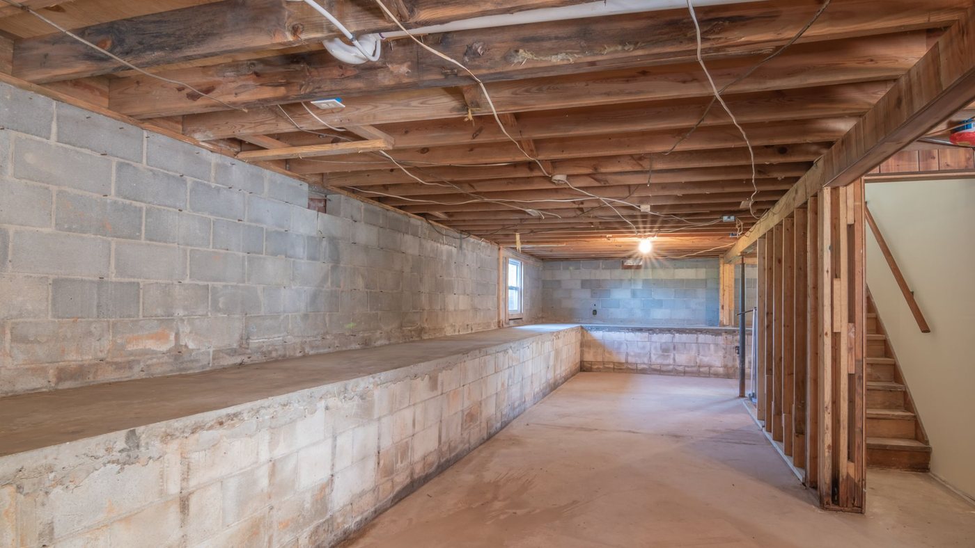 Unfinished basement with bare concrete block foundation walls, exposed floor joists and rim joists overhead, and a framed partition beside the stairs