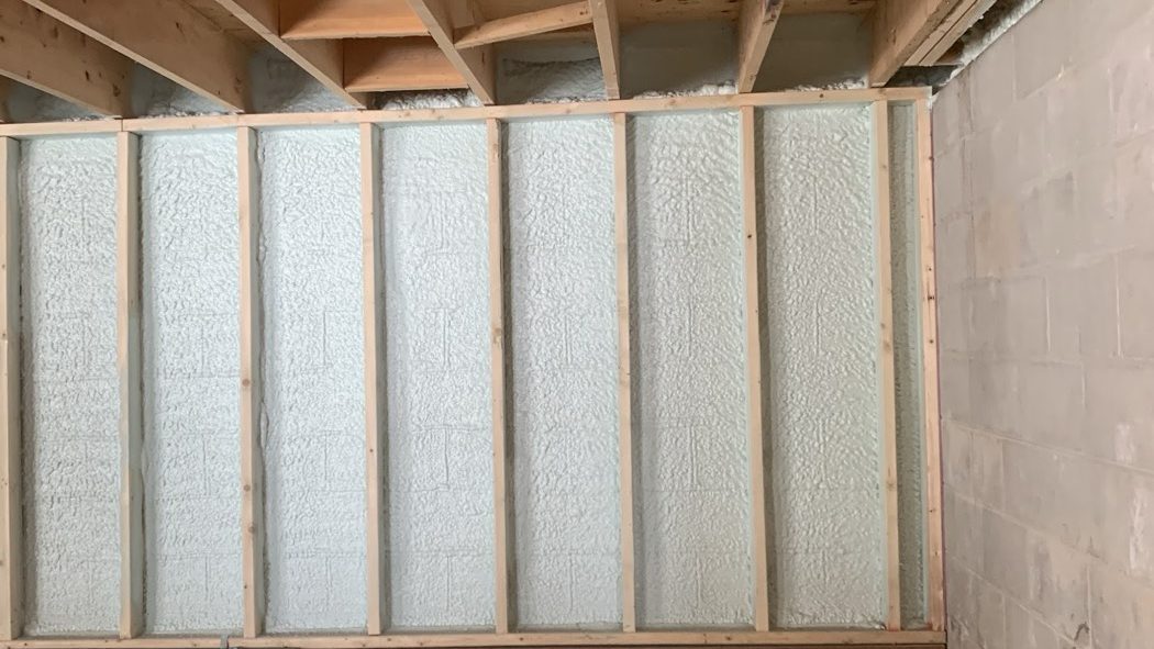 Framed basement wall with closed cell spray foam filling the bays between the studs, against a concrete block foundation, with the floor joists and rim area above