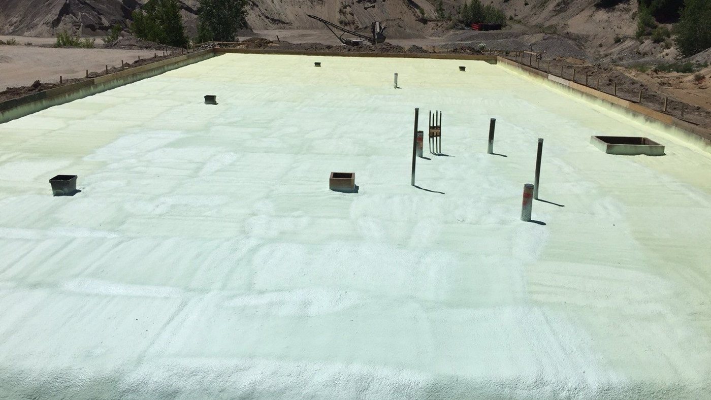 Spray foam sprayed to an even surface over a shop floor before the pour, drains and plumbing stubs standing through it: the same penetrations a vapour barrier basement slab has to seal